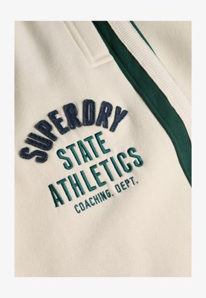 Tissu crème avec du texte brodé : "SUPERDRY STATE ATHLETICS COACHING DEPT." en marine et turquoise, avec des accents côtelés verts.