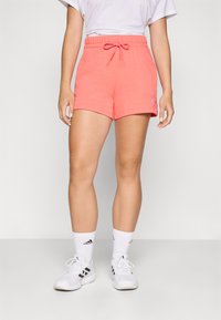 Calções de algodão corais com cintura elástica e cordão; apresentam um pequeno logo na bainha; combinados com meias e sapatos brancos.
