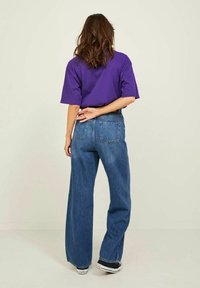 Žena s dlouhými vlnitými hnědými vlasy stojí zády, má na sobě volné fialové tričko, široké modré džíny a černé tenisky.