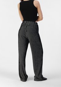 Femme portant un haut sans manches noir et un pantalon noir ample avec des rayures verticales blanches, se tenant les bras croisés, vue de dos.