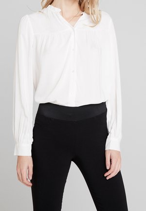 Kobieta w białej bluzce zapinanej na guziki z długimi rękawami i czarnych spodniach z wysokim stanem na jednolitym szarym tle.