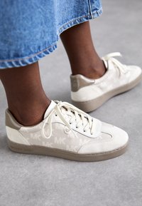 Baskets en tissu blanc avec des accents beiges, présentant un motif répétitif du logo MK, une semelle en caoutchouc et des lacets plats.