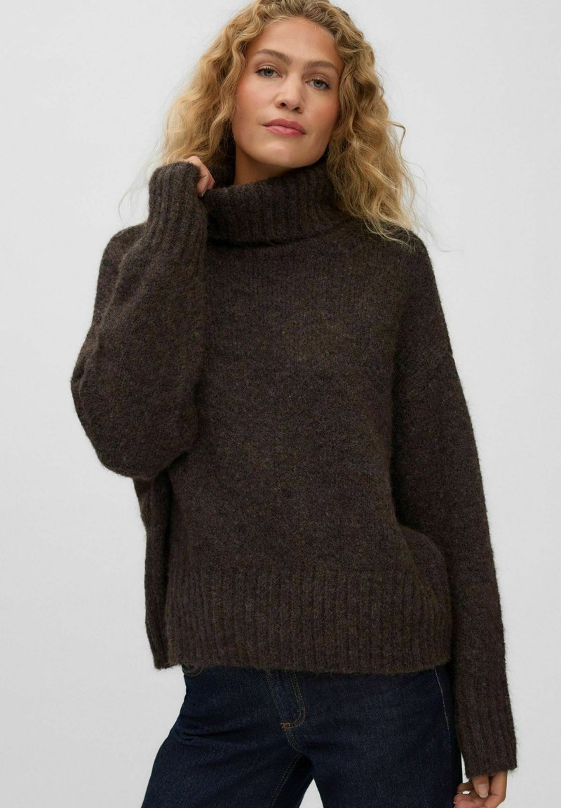 Femme aux cheveux blonds bouclés portant un pull en maille épaisse à col roulé marron foncé et un jean bleu foncé sur un fond uni.