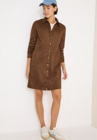 Vestito camicia in suede marrone con maniche lunghe, chiusura a bottoni davanti e due tasche anteriori. Abbinato a sneakers chiare e un cappellino blu.