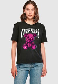 Sort T-shirt med korte ærmer i sort, prydet med et cartoon-illustration af en pink teddybjørn med X-formede øjne og teksten "PRØV AT HÅNDTERE AL DENNE SØDME" ovenover.