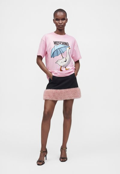 Γυναίκα που φοράει ένα ροζ t-shirt Moschino με σχέδιο χήνας και ομπρέλας, μια μαύρη φούστα με ροζ γούνινη επένδυση και μαύρα παπούτσια με λουράκι στον αστράγαλο.