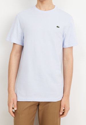 Osoba nosząca gładki biały T-shirt z krótkim rękawem, z małym zielonym logo krokodyla na piersi oraz brązowe spodnie.