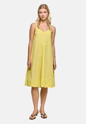 Woman stands facing forward, wearing a loose yellow sleeveless dress with lace trim and scalloped hem, paired with black heeled sandals.
