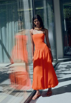 Robe maxi sans manches orange avec un corsage ajusté, une jupe évasée et un tissu texturé. Portée avec un collier en perles volumineux et des sandales plates.
