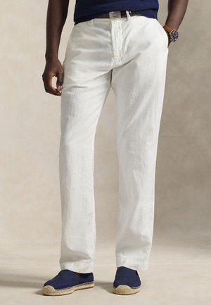 Bărbat purtând pantaloni albi, espadrile bleumarin, o cămașă bleumarin, un ceas maro din piele și o curea neagră cu cataramă argintie.