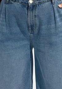 Jasnoniebieskie dżinsy o luźnym kroju, z plisami w pasie i klasycznym zapięciem na guziki. Teksturowana tkanina z widocznym przeszyciem.