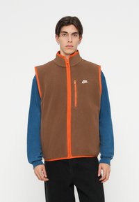 Hnědá fleecová vesta s vysokým límcem, oranžovým zipem a lemem. Obsahuje přední kapsu na zip a logo Nike na hrudi. Nositá přes modré dlouhé rukávy.