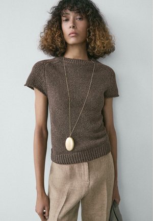 Giovane donna con capelli ricci che indossa una maglia marrone a maniche corte in maglia, pantaloni beige e una lunga collana con ciondolo ovale.