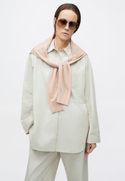 Chemise boutonnée oversize gris clair avec une poche poitrine, associée à un pull rose noué autour du cou. Lunettes de soleil marron oversize.