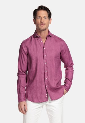 Man draagt een magenta overhemd met lange mouwen en witte broek, staat met één hand in de zak tegen een effen achtergrond.