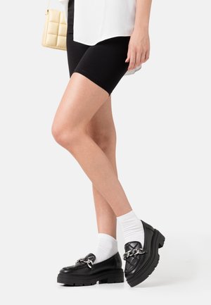 Black chunky loafers with a silver chain accent, paired with white ankle socks and black cycling shorts. Smooth material and durable sole.