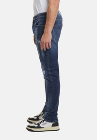 Hombre vistiendo jeans ajustados de mezclilla oscura desgastada y zapatillas negras y blancas, de pie de lado contra un fondo simple.