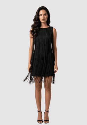 Mujer con cabello largo y oscuro, vestida con un mini vestido negro sin mangas con flecos y tacones negros con tiras en el tobillo, de pie frente a un fondo claro y liso.