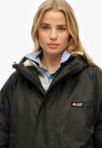 Chaqueta negra impermeable con capucha, que presenta un frente con cremallera y un parche de logotipo. Llevada sobre una camisa de cuello azul con acentos grises.