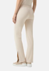 Pantalons stretch beiges avec une coupe slim, présentant des fentes sur les ourlets et une texture lisse. Portés avec des baskets de couleur claire.