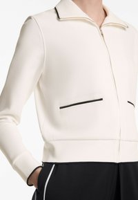 White zip-up jacket with a folded collar, black accent piping on pockets, and ribbed cuffs. Smooth texture, fitted design.