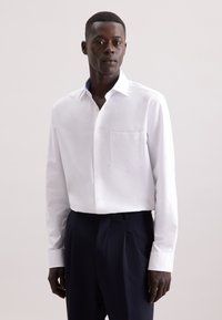 Camicia bianca a maniche lunghe in tessuto morbido con taschino sul petto e colletto interno a fantasia, abbinata a pantaloni scuri. Design semplice e sartoriale.