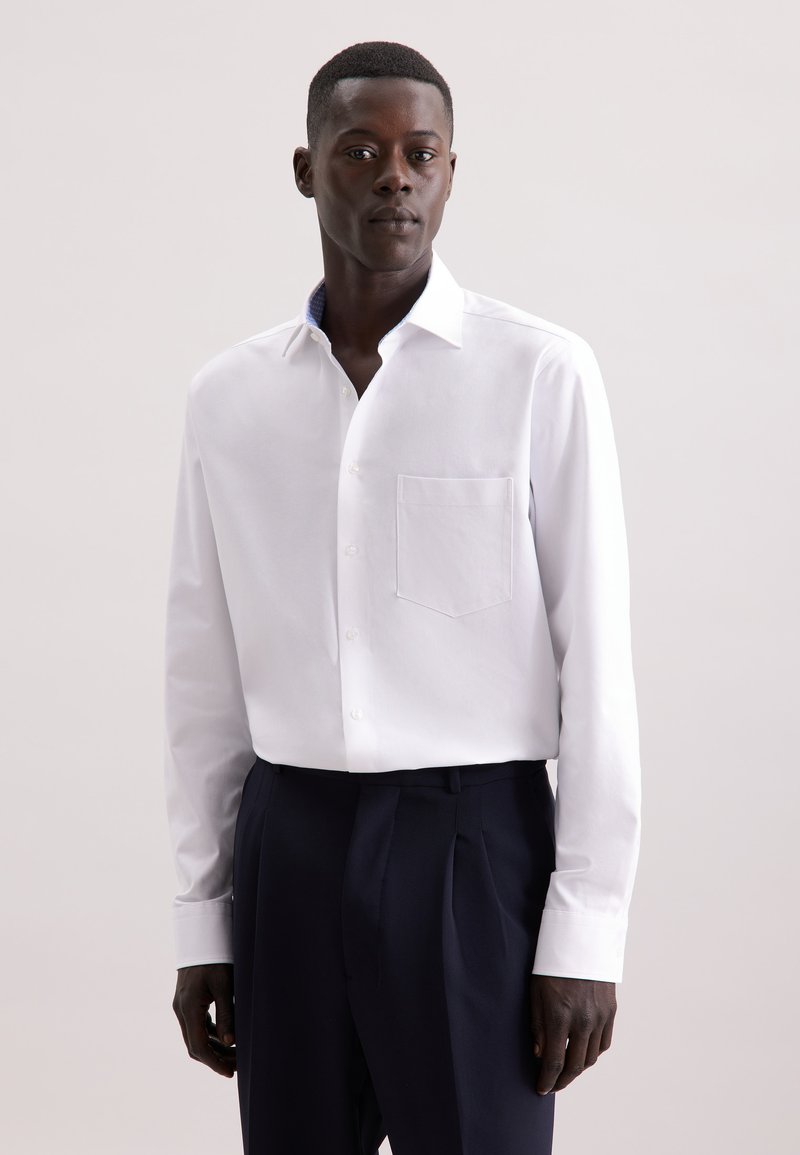 Camicia bianca a maniche lunghe in tessuto morbido con taschino sul petto e colletto interno a fantasia, abbinata a pantaloni scuri. Design semplice e sartoriale.