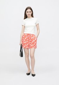 Bílé tričko s krátkým rukávem spojené s oranžovými šortkami s květinovým vzorem a černými balerínkami. Modelka drží malou černou kabelku.