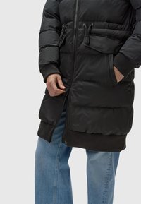 Black puffer coat with a zip front, elastic cuffs, adjustable drawstring waist, and ribbed hem, paired with light blue wide-leg jeans.