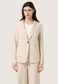 Blazer beige su misura realizzato in tessuto morbido. Presenta una chiusura a un bottone e tasche laterali. Spalle strutturate e design classico dei revers.