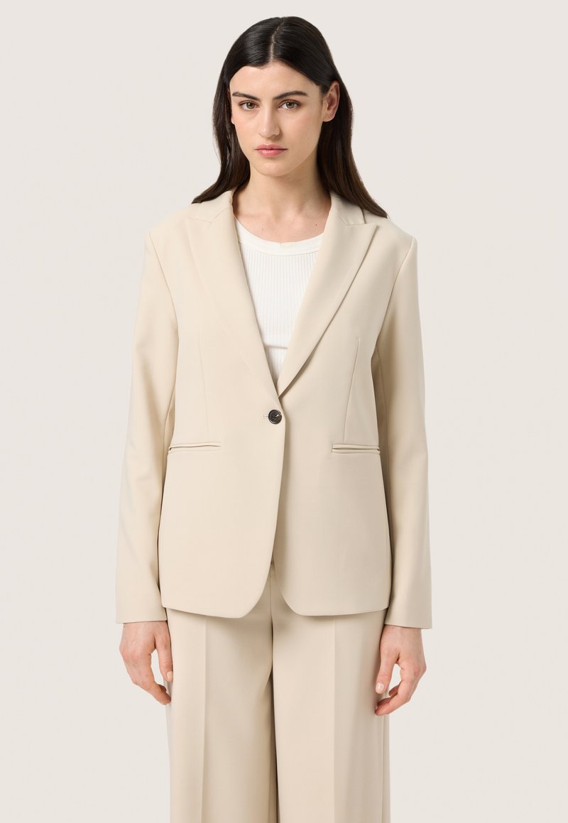 Blazer beige su misura realizzato in tessuto morbido. Presenta una chiusura a un bottone e tasche laterali. Spalle strutturate e design classico dei revers.