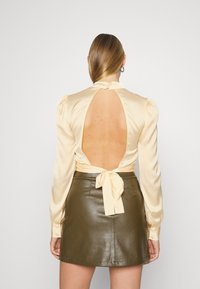 Blusa de cetim em amarelo pálido com um decote profundo nas costas, mangas longas e punhos com botões, combinada com uma saia justa de couro verde-oliva.