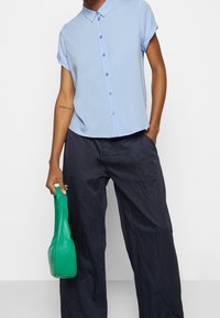 Chemise bleu clair à col avec manches courtes retroussées, boutonnée devant, associée à un pantalon large bleu marine foncé et un sac à main vert texturé.