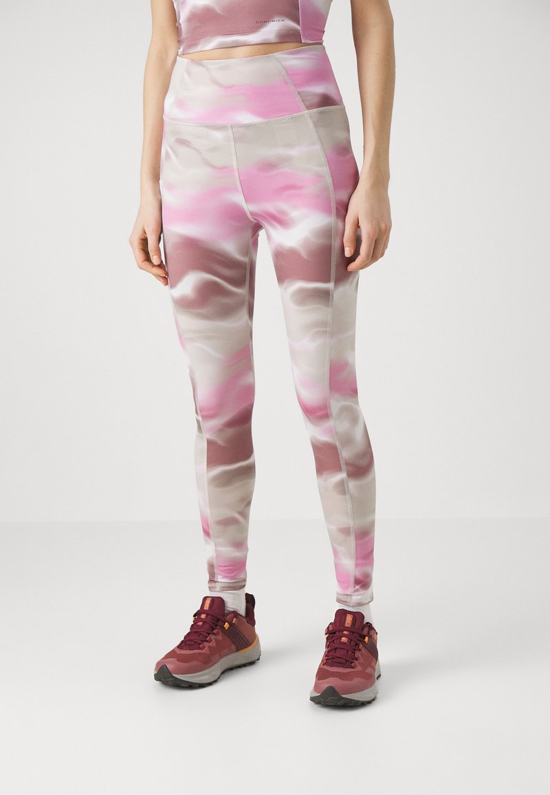 Kvinna som bär rosa, beige och vita mönstrade leggings med en matchande kort tröja och vinröda löparskor, stående på ett vitt golv.