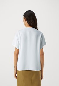Blusa azzurra a maniche corte con una texture liscia, collo rotondo e discrete cuciture sul retro, abbinata a una gonna marrone.