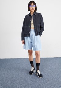 Veste en denim noir, shorts en denim bleu clair, chaussettes noires et baskets noires. Modèle portant des lunettes de soleil bleues transparentes. Fond sobre.