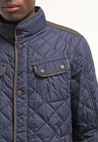 Marineblå quiltet jakke med høj krave, der har to brystlommer med knapklapper, kontrasterende tan-detaljer og et glat, tekstureret stof.