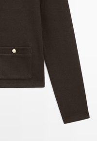 Dark brown long sleeve garment with a small pocket on the left side, featuring a gold-tone button on the pocket flap.