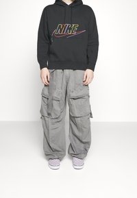 Black hoodie with colorful Nike logo, gray cargo pants with multiple pockets, and purple checkered slip-on shoes. Overall relaxed fit.