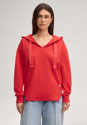 Kobieta nosząca luźną czerwoną bluzę z szerokimi sznurkami i jasnoniebieskie dżinsy, stojąca na tle jednolitego, jasnego tła.