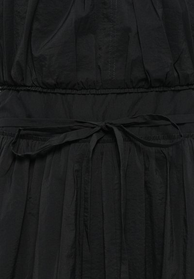 Rochie neagră dintr-un material ușor, texturat; prezintă un talie elastică strânsă cu o fundiță și o fustă plisată.