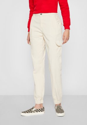 Persona vistiendo pantalones cargo color crema con puños elásticos, una camiseta roja de manga larga y zapatos tipo slip-on a cuadros en blanco y negro.