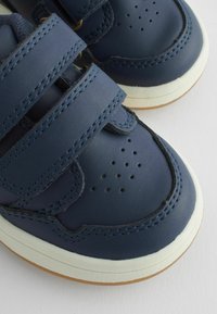 Chaussures en cuir bleu marine avec deux sangles Velcro ajustables, design de pointe perforé, semelle en caoutchouc blanche et semelle extérieure texturée marron.