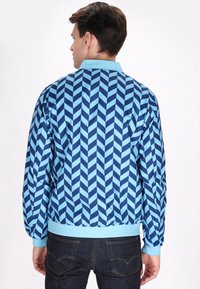 Giacca bomber imbottita con un audace motivo a zigzag blu, polsini e orlo a coste, e un colletto azzurro. Finitura liscia e lucida.