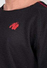 Zwarte sweatshirt met textuur, voorzien van een geborduurd rood gorillalogo op de borst en subtiele rode accentstiksels langs de naad.