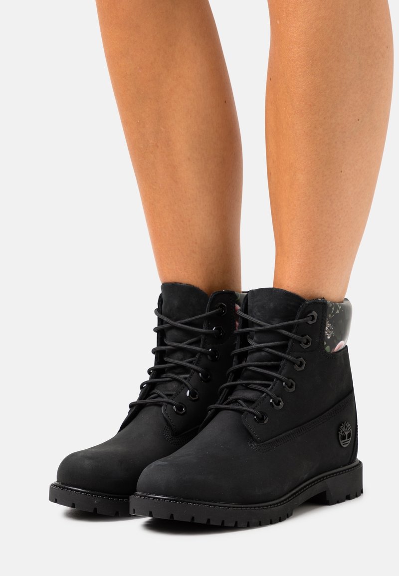 Bottines noires à lacets avec semelles robustes portées sur jambes nues, avec des cols rembourrés et des accents à motif floral.