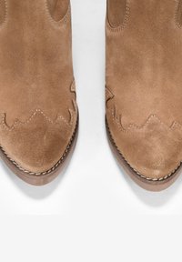 Botas de tobillo de ante marrón con punta afilada, con costuras en zigzag a lo largo del borde superior y una delgada suela de cuero. Acabado texturizado.