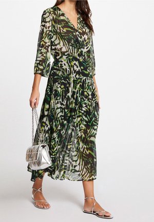 Kvinde iført en grøn og sort mønstret midikjole med gennemsigtige ærmer, holder en sølvfarvet quiltet håndtaske og har sølvfarvede sandaler med stropper på.