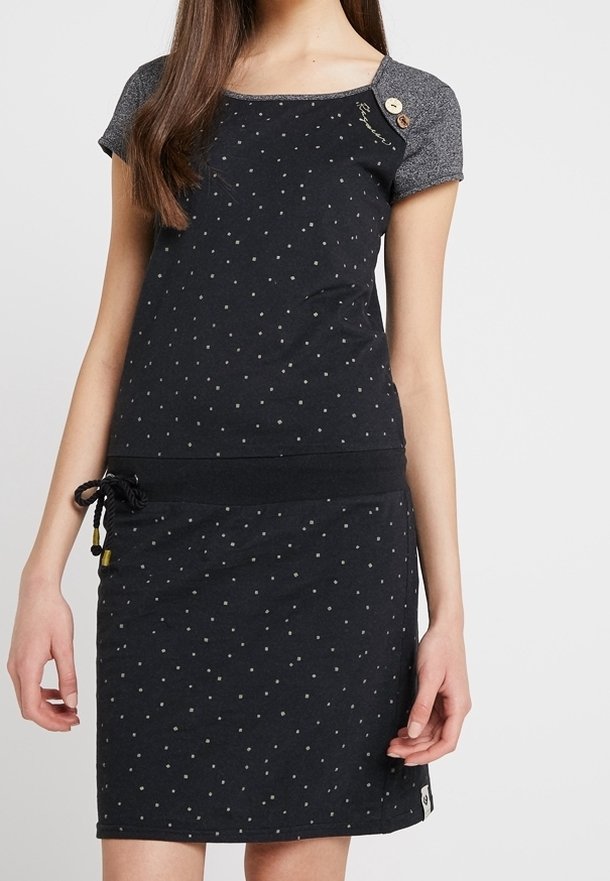 Femme portant une robe noire à manches courtes avec de petits pois blancs, des manches grises et un détail à cordon sur le côté gauche de la taille.