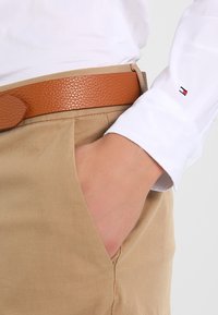 Beige Baumwollhosen mit einem braunen, strukturierten Ledergürtel. Weißes Langarmhemd mit einem kleinen Logo, das am Bündchen sichtbar ist.